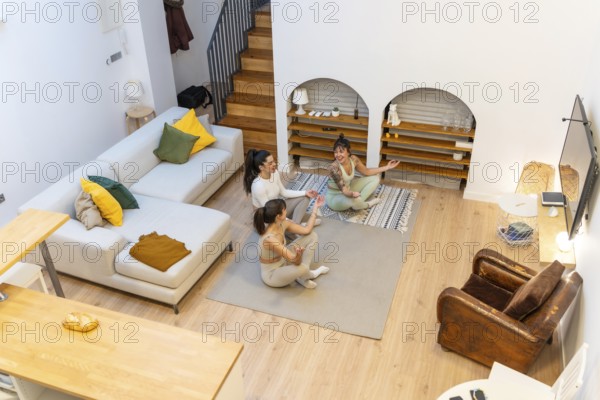 Three young women sitting in a living room, smiling and relaxing on yoga mats, enjoying a peaceful moment of wellness and mindfulness during their fitness workout routine