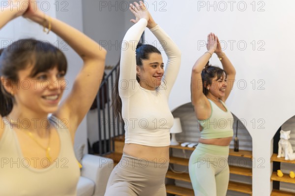 Three diverse women practicing yoga together at home, smiling and balancing in a sunlit modern apartment, promoting fitness, mindfulness, friendship and healthy lifestyle