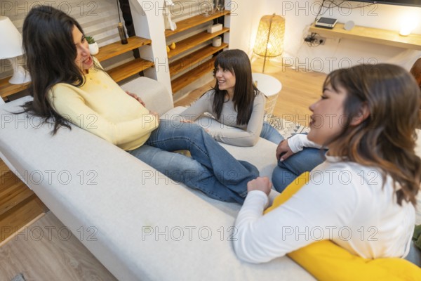 Pregnant woman relaxing on a sofa with two close friends in a cozy living room, sharing laughter, support and anticipation of motherhood during a warm, casual visit