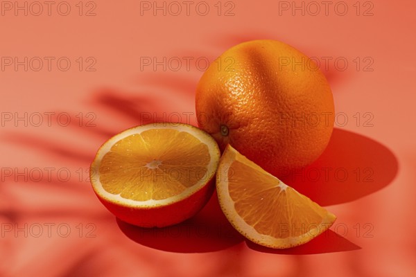 On the coral surface, next to two neatly sliced slices, there is a whole orange. Sunlight casts soft shadows, emphasizing the freshness and bright color of the fruit