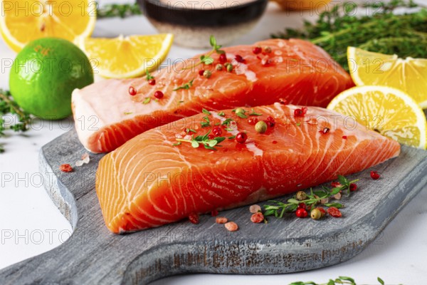 Two pieces of salmon fillet sit on a wooden cutting board. They are topped with herbs and pink peppercorns. Slices of lemon and lime surround the fish, creating a vibrant setting for cooking