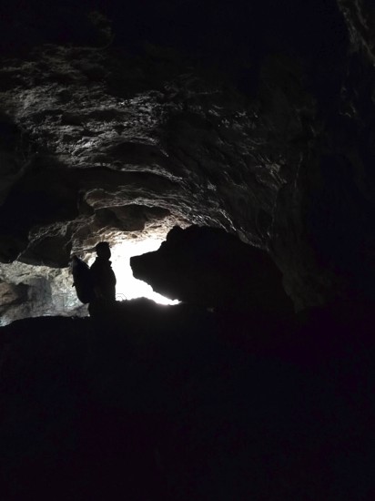 Silhouette of a person in a dark cave entrance with a ray of light from outside and a rock that looks like a big boar (aprum), Franconian Switzerland, Germany