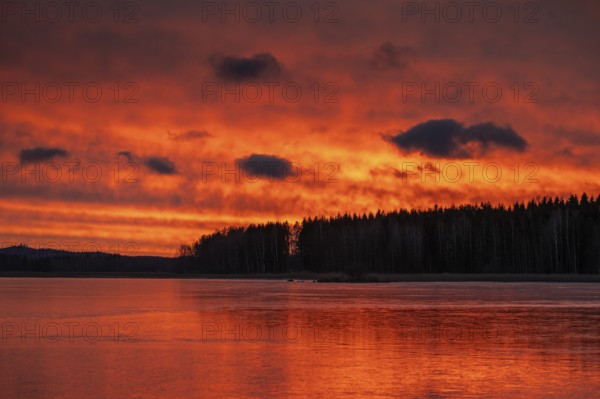A dramatic sunset over a still, frozen lake and forest with orange-red hues in a cloudy sky, Finland