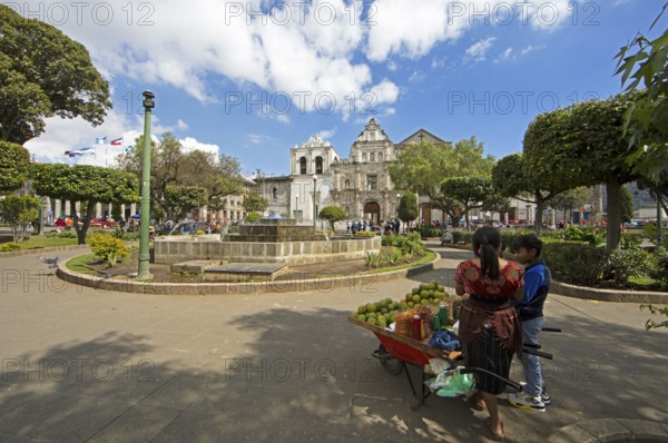 Parque Centroamérica or Parque Central, a Mayan woman in traditional dress selling oranges, in the back the cathedral or Catedral del Espíritu Santo, Quetzaltenango or Xela, Quetzaltenango province, Guatemala