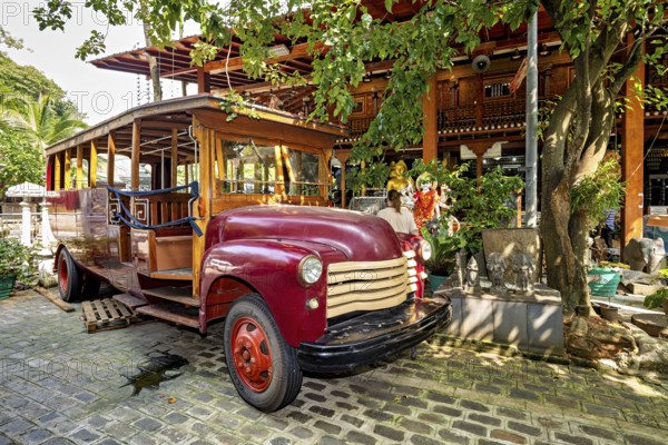 Red antique vehicle in front of a wooden building in tropical surroundings, surrounded by trees, nostalgic atmosphere, vintage car in the temples of Colombo in Sri Lanka