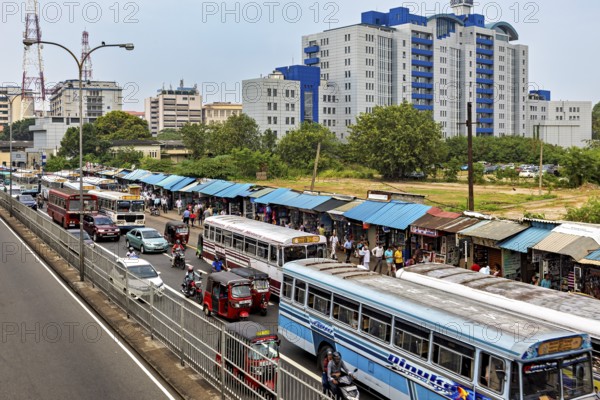 City street with several buses and market stalls, surrounded by large buildings and trees, buses in traffic from Colombo in Sri Lanka