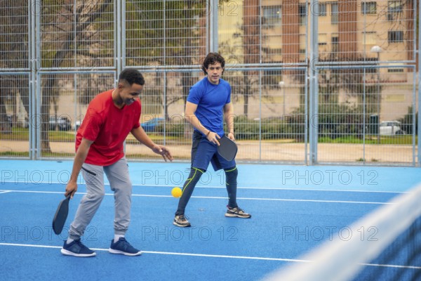 Two diverse young men compete on a blue outdoor pickleball court, focused and energetic as they rally with paddles under sunny urban skies, enjoying sport and fitness together