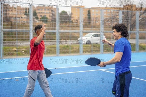 Two men in red and blue shirts smile and high five on a vibrant blue outdoor pickleball court, celebrating teamwork, friendship and active, joyful recreation