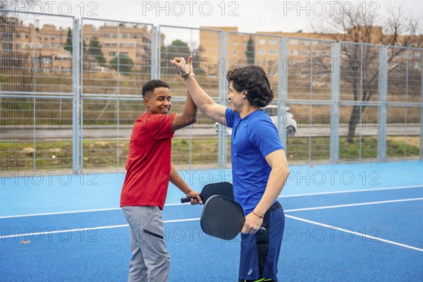 Two smiling male athletes, one african american and one caucasian, giving a high five on a blue outdoor pickleball court, celebrating teamwork and good sportsmanship after a game