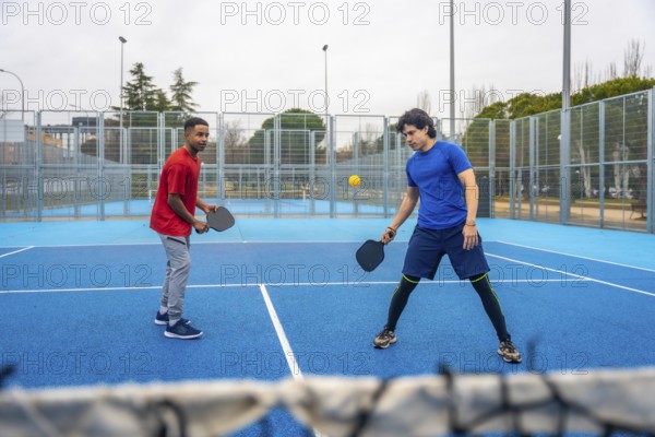 Two young men playing pickleball, hitting the ball over the net during a competitive match on an outdoor blue court, demonstrating focus and active lifestyle