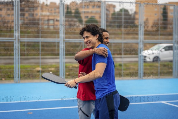 Two diverse male friends smiling and hugging on a blue pickleball court after a game, demonstrating companionship, teamwork, and healthy competition in an urban setting