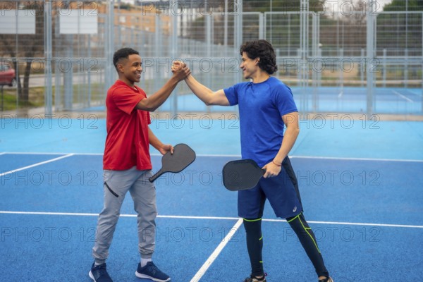 Two smiling male pickleball players of different ethnicities shaking hands on a vibrant blue court after a game, celebrating teamwork and sportsmanship