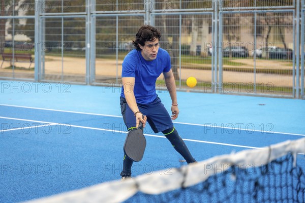 Young man concentrating on a pickleball match on a blue outdoor court, holding his paddle ready to hit the yellow ball over the net, focused and athletic in action