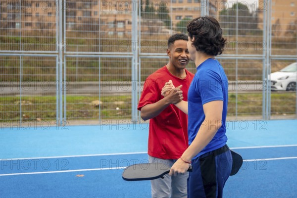 Two young multi ethnic men shaking hands and smiling on a blue pickleball court after a match, showing sportsmanship, friendship and active outdoor recreation