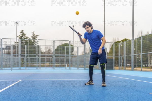 Young athletic man wearing blue t shirt and shorts focusing on game, hitting a yellow ball with a paddle during a pickleball match on a blue outdoor court