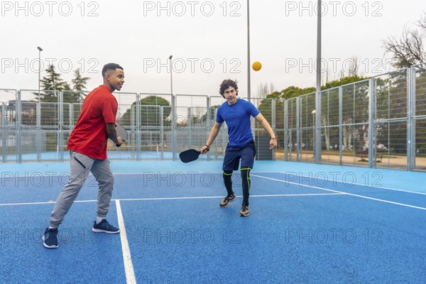 Two young menone african american, one caucasiancompete in an energetic outdoor pickleball rally, paddles in hand on a bright blue court, showcasing fitness and friendship