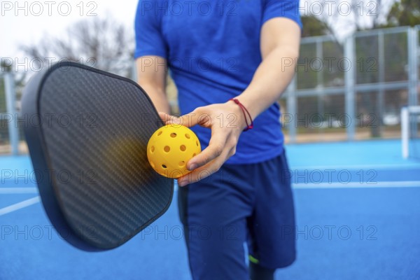 Athlete preparing for a game, holding a perforated yellow pickleball and a black paddle on a blue court, representing a popular racquet sport and active lifestyle