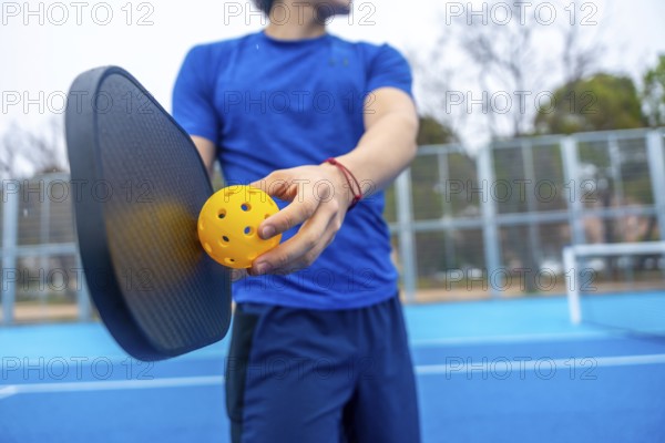 Athlete preparing to serve, holding a yellow pickleball and paddle on a vibrant blue outdoor court, ready for a recreational game or competitive match