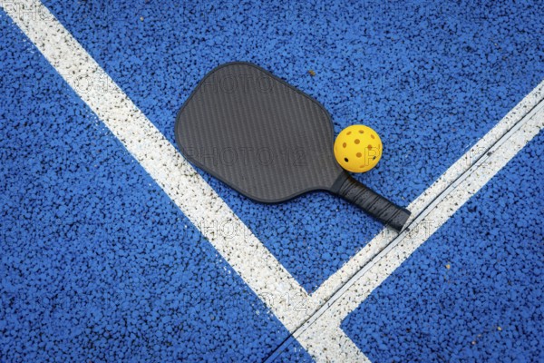 Pickleball paddle and yellow wiffle ball resting on the vibrant blue court surface with distinct white boundary lines, representing an active lifestyle and recreational sport