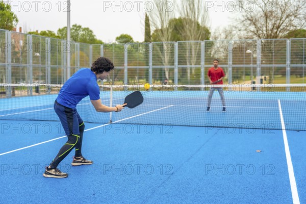 Two young men actively engaging in a game of pickleball, one hitting the yellow ball with a paddle, on a bright blue outdoor court with a net, enjoying leisure and fitness