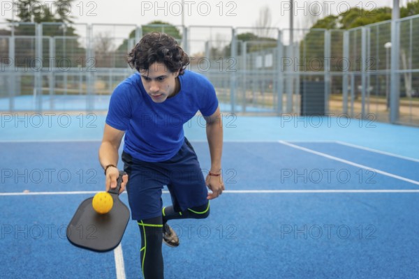 Male athlete concentrating while holding a pickleball paddle and yellow ball, ready to serve or return on a vibrant blue outdoor court, engaging in active sport and fitness