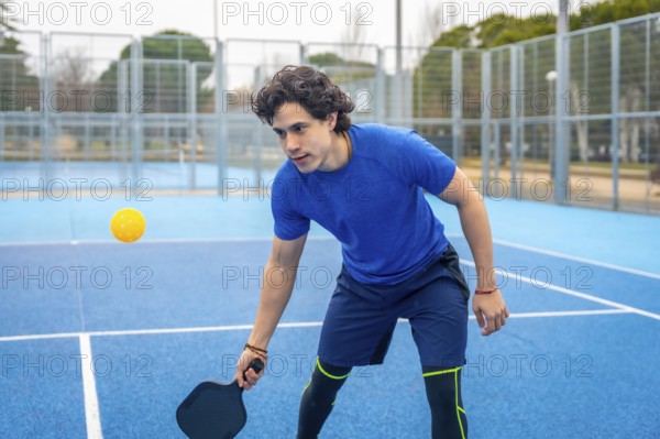 Young man holding a pickleball paddle, focused and ready to hit the yellow ball during an outdoor sports game on a vibrant blue court, representing fitness and recreation