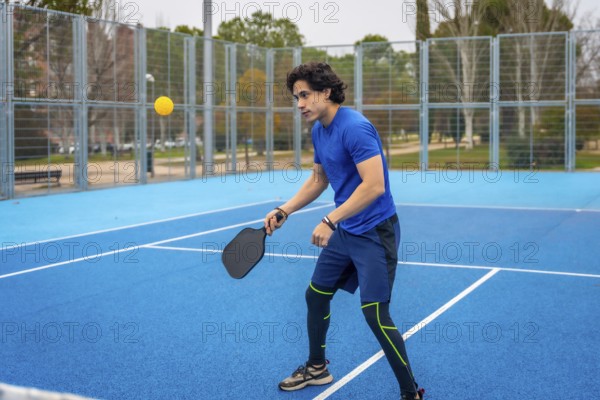 Young man playing pickleball on a blue outdoor court, striking a yellow ball with a black paddle, showing athletic movement, focus and active leisure lifestyle in sportswear