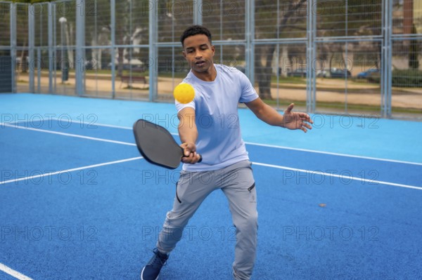 Young man strikes a yellow pickleball with a paddle on a vibrant blue outdoor court, showcasing energetic movement, focus, and active summer leisure in modern recreational sport