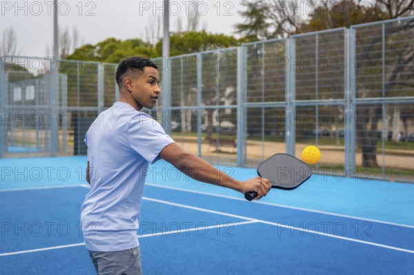 Young man wearing a light blue t shirt and grey shorts holding a paddle, hitting a yellow pickleball on a vibrant blue outdoor court during a match, focusing on the game