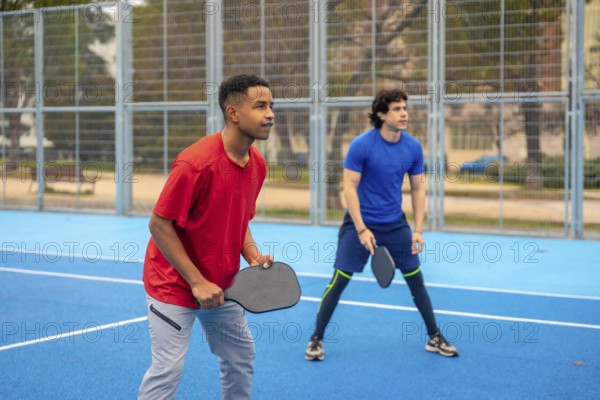Two young diverse men holding paddles and concentrating on the game, actively participating in a pickleball match on a vibrant blue court surrounded by a fence and trees