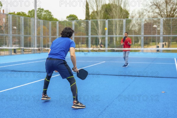 Two male athletes playing an intense pickleball game on a vibrant blue court, holding paddles and focusing on hitting the ball over the net during a competitive and active sporting event outdoors