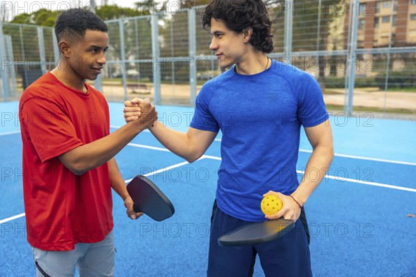 Two young diverse male athletes shaking hands on a blue pickleball court, holding paddles and a pickleball, symbolizing fair play, competition, and positive human connection after a game