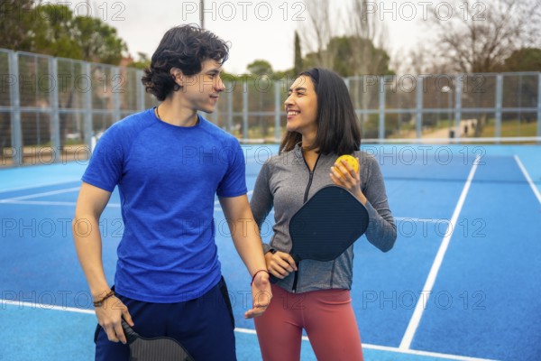 Young man and woman standing together on a vibrant blue pickleball court, holding paddles and ball, sharing a happy moment before playing the game outdoors