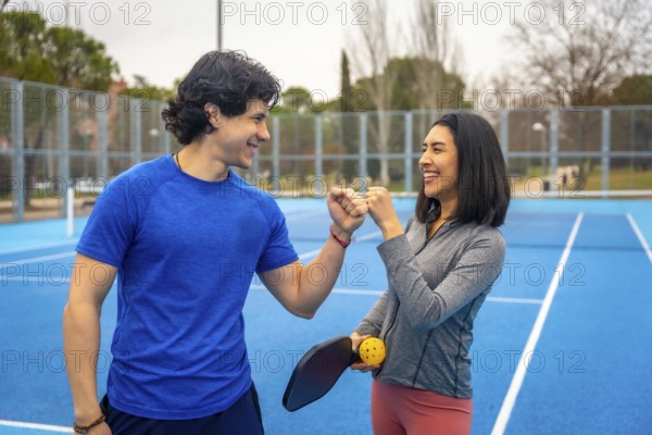 Young man and woman celebrating success with a fist bump, smiling and standing together on a bright blue pickleball court while holding a paddle and ball, representing teamwork and active lifestyle