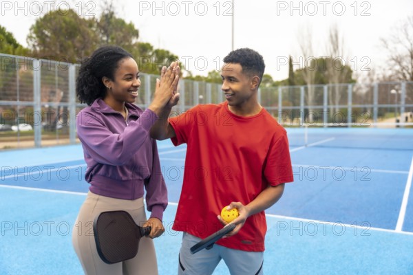 Young diverse couple playing pickleball on a blue court, celebrating with a high five, showing teamwork, friendship, and positive energy during outdoor exercise