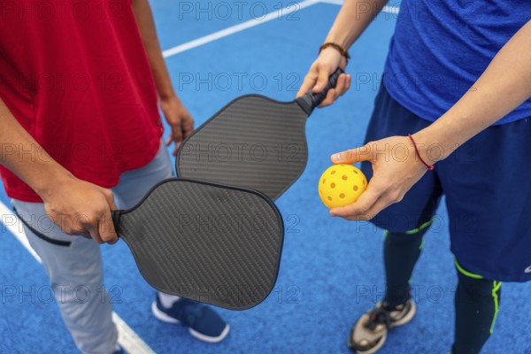 Two people are ready to play pickleball, holding paddles and a bright yellow wiffle ball on a vibrant blue court, highlighting recreation and active lifestyle