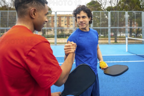 Two diverse male pickleball players shaking hands on a blue court, proudly displaying their paddles and ball, celebrating fair play and friendship after a game