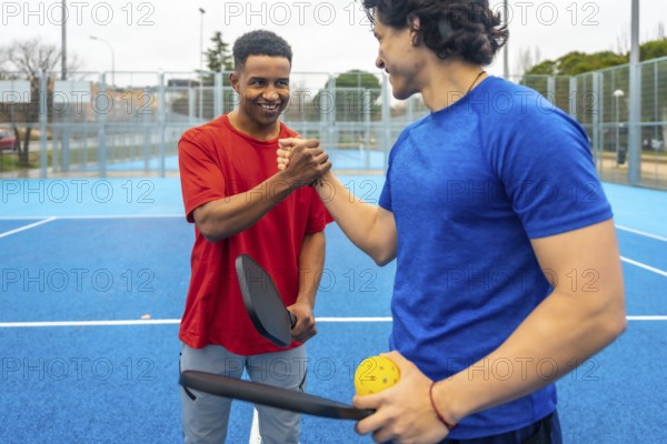 Two male pickleball players shaking hands on a blue outdoor court after a match, showing sportsmanship, friendship and teamwork in a casual, active urban summer setting