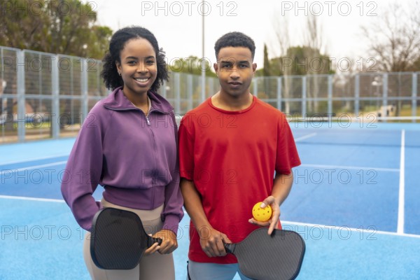 Young couple standing on a blue pickleball court, holding paddles and a ball, smiling at the camera, representing an active lifestyle and healthy sport