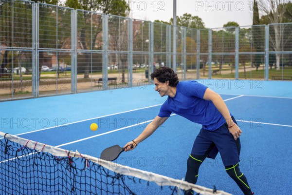 Young man in sports clothing enthusiastically playing pickleball on a bright blue court, hitting a yellow ball with a paddle, enjoying the active sport outdoors