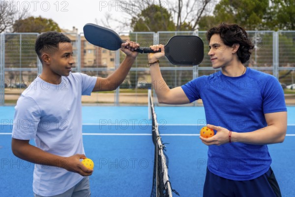 Two men stand on an outdoor pickleball court holding paddles and balls, sharing a friendly moment that highlights teamwork, competition, and an active lifestyle