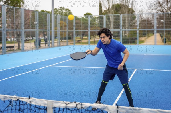 Young man in blue activewear playing pickleball on a blue court, intently hitting a yellow ball with a paddle, enjoying the modern racket sport outdoors