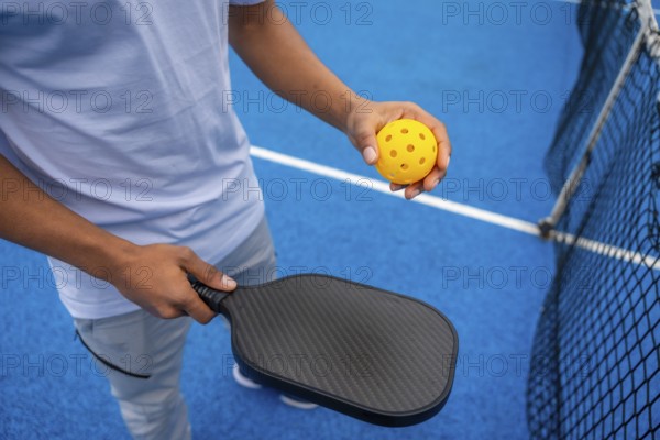 Person standing on a bright blue pickleball court, holding a black paddle and a yellow perforated pickleball, ready to play and engage in competitive sport and active lifestyle