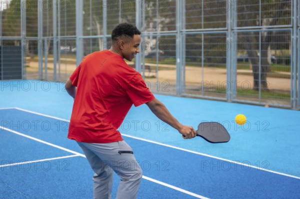 Young athletic man in sportswear striking a pickleball with a paddle on a vibrant blue outdoor court, focused and smiling while enjoying energetic summer play and fitness