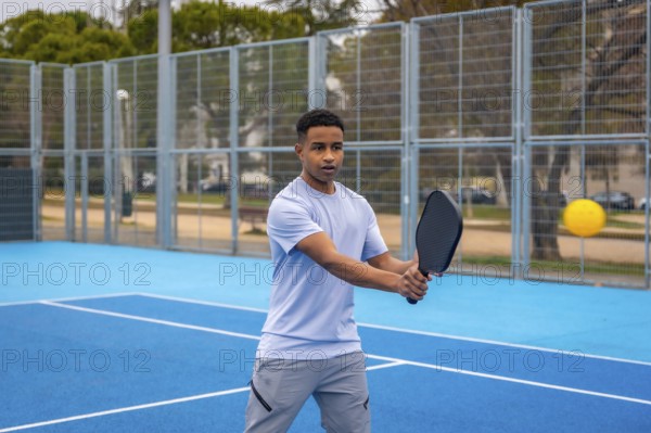Young man poised to strike a yellow ball with a paddle on an outdoor blue pickleball court, focused and athletic during a competitive match on a sunny day