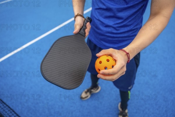 Player standing on a vibrant blue pickleball court, holding a textured paddle and an orange wiffle ball, preparing to start a competitive game or practice session