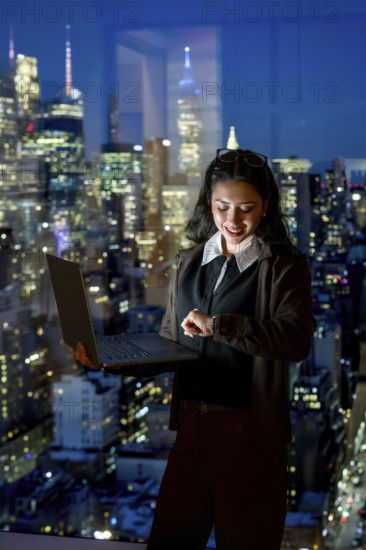 Young businesswoman standing by a large window, working on a laptop and checking her smartwatch while cityscape lights illuminate the background at a late hour, representing dedication and success