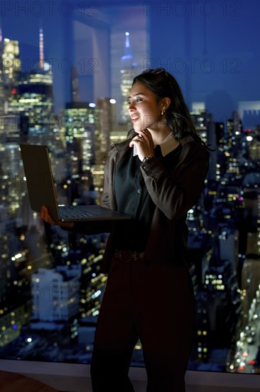 Young businesswoman standing confidently by a large window with her laptop, looking out at the illuminated manhattan city skyline at night, representing professional success and urban achievement