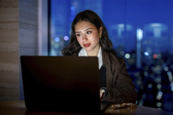 Young businesswoman focusing on her laptop screen, working late hours in a modern office overlooking the illuminated cityscape of manhattan, demonstrating dedication and ambition