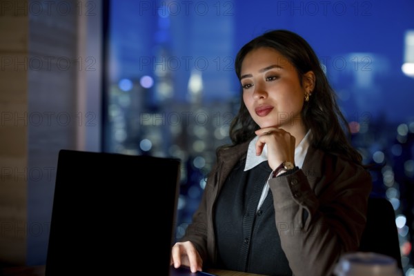 Young businesswoman with elegant hairstyle working late hours, researching or analyzing data on her laptop, overseeing the illuminated manhattan city skyline from her modern office